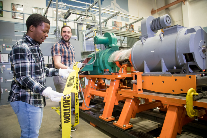 Ph.D. student Thabiso Mabote, left, and College of Engineering faculty member Eduardo Cotilla-Sanchez put caution tape around a machine testbed. Machine testbeds are useful to validate machine dynamics in a time-domain simulation such as was used in the smart meter research. (photo provided by Eduardo Cotilla-Sanchez)