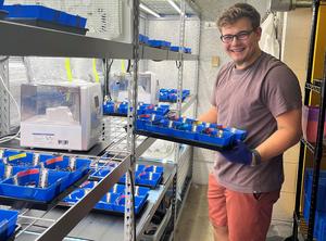 Liam Reynolds is holding a tray with blue containers next to metal shelves with similar trays.