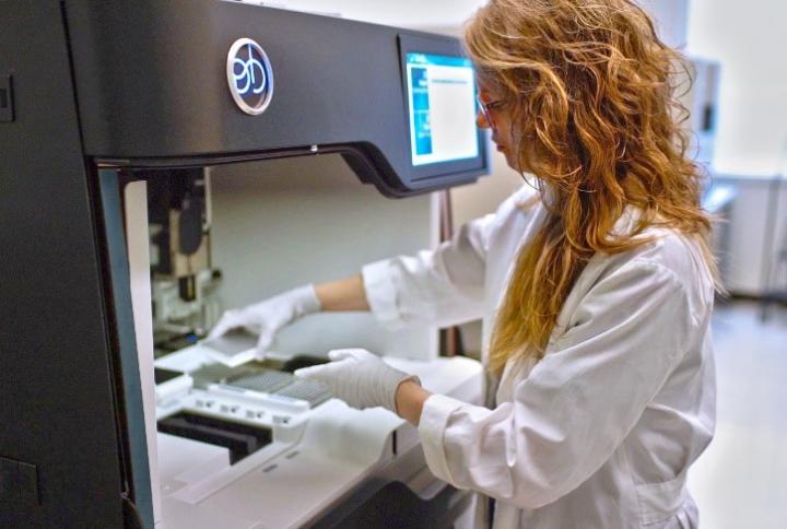 A Young Woman in a Lab Coat and Gloves Places a Small Tray of Biosamples in a Machine for Analysis