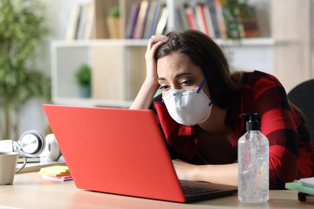 Bored student woman with mask looking at laptop sitting on a desk in coronavirus confinement at home