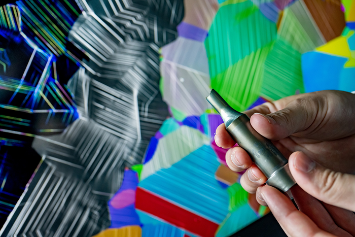 In the foreground, a researcher holds a peice of aircraft engine alloy broken in one of their experiments. In the background is a compliation of images taken with a scanning electron microsope and a show a visual representation of atomic slip -- the nanoscale mechanism behind metal failure.