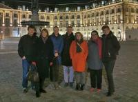 Seven people in winter coats in the evening in a European square with lit building in background.