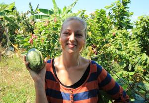 female researcher holding green, oval cocoa pod, standing in landscape